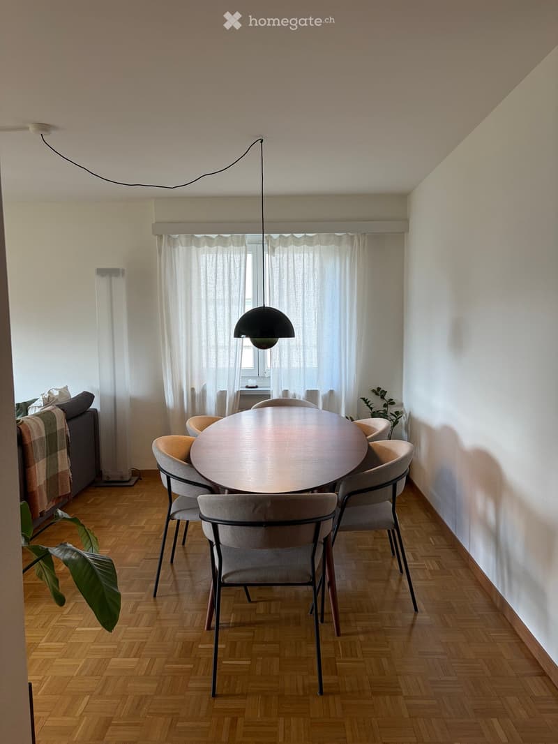 Dining area with a round wooden table surrounded by six chairs, a sofa with a blanket, a floor lamp, and a pendant light hanging from the ceiling. The room has wooden flooring and white walls with large windows covered by sheer curtains.