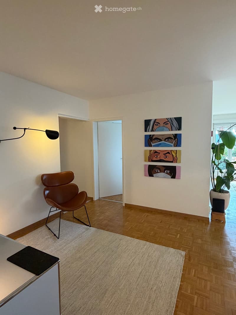 A modern, minimalist room with wooden flooring, a brown leather chair, a small table with a laptop, a wall-mounted lamp, and several pieces of artwork on the wall. A large potted plant is near the window.