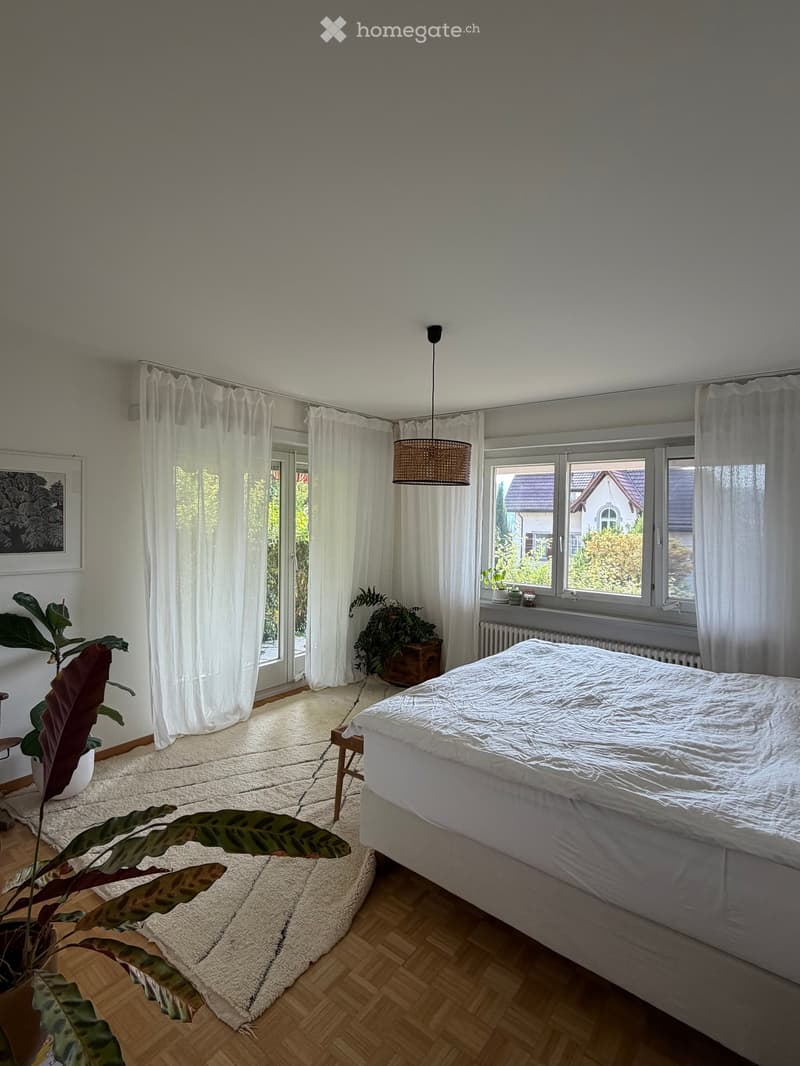 Bright bedroom with wooden flooring, large windows with white curtains, a bed with white bedding, potted plants, and a hanging lamp.