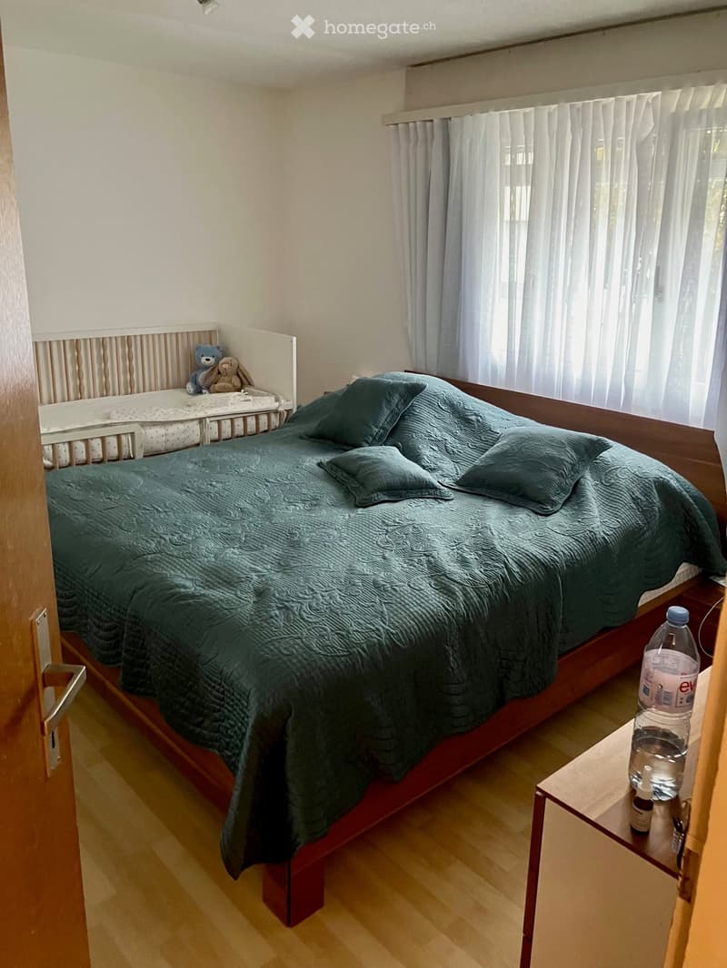 A cozy bedroom with a wooden bed, green bedding, pillows, a nightstand with a water bottle, and a small toy on the bed.
