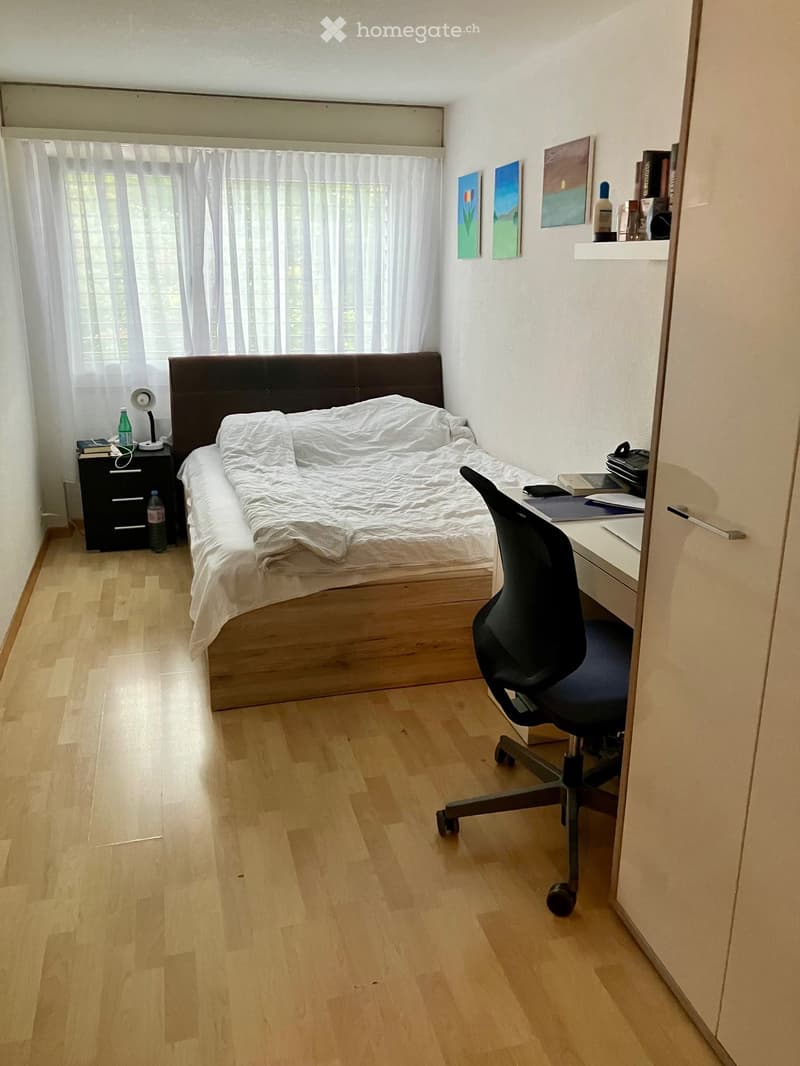 A small, well-lit bedroom with a wooden floor, a bed with white bedding, a nightstand, a desk with a chair, and several books and decorative items on shelves.