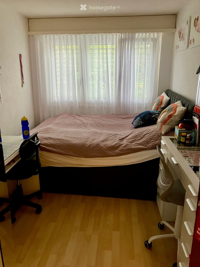 A cozy bedroom with a bed, desk, and chair. The bed has a pink quilt and colorful pillows, and the room features wooden flooring and white curtains on the window.