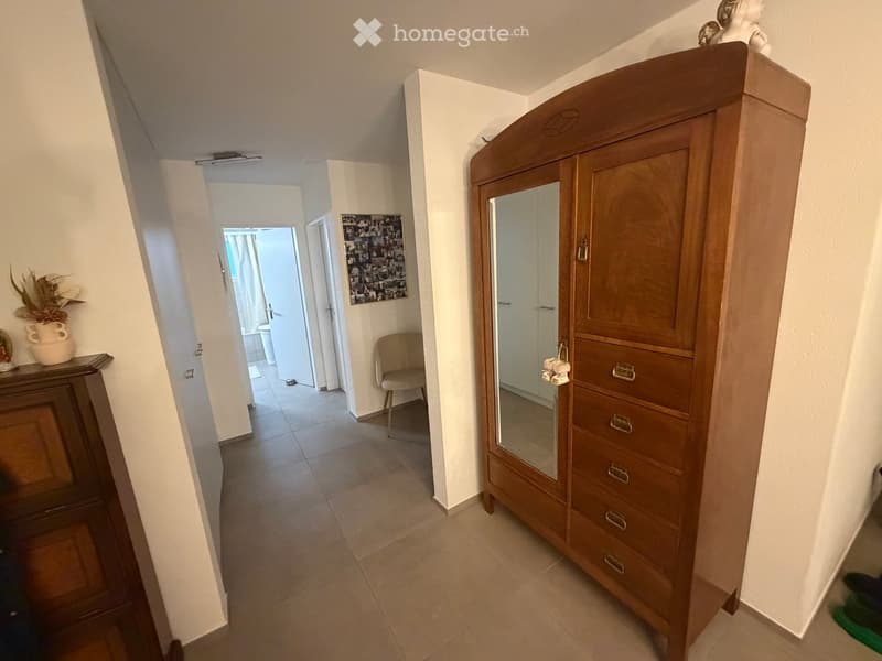 Hallway with tiled floor, wooden wardrobe with mirrored doors, small table with vase, chair, and a photo collage on the wall.