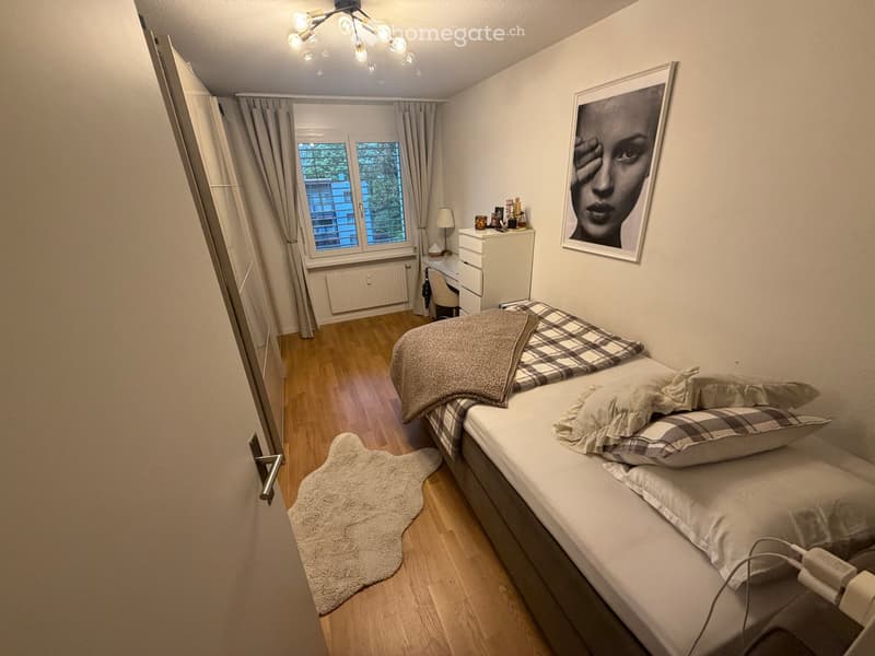 Cozy bedroom with a single bed, pillows, blanket, nightstand, lamp, picture frame, and a window with curtains.