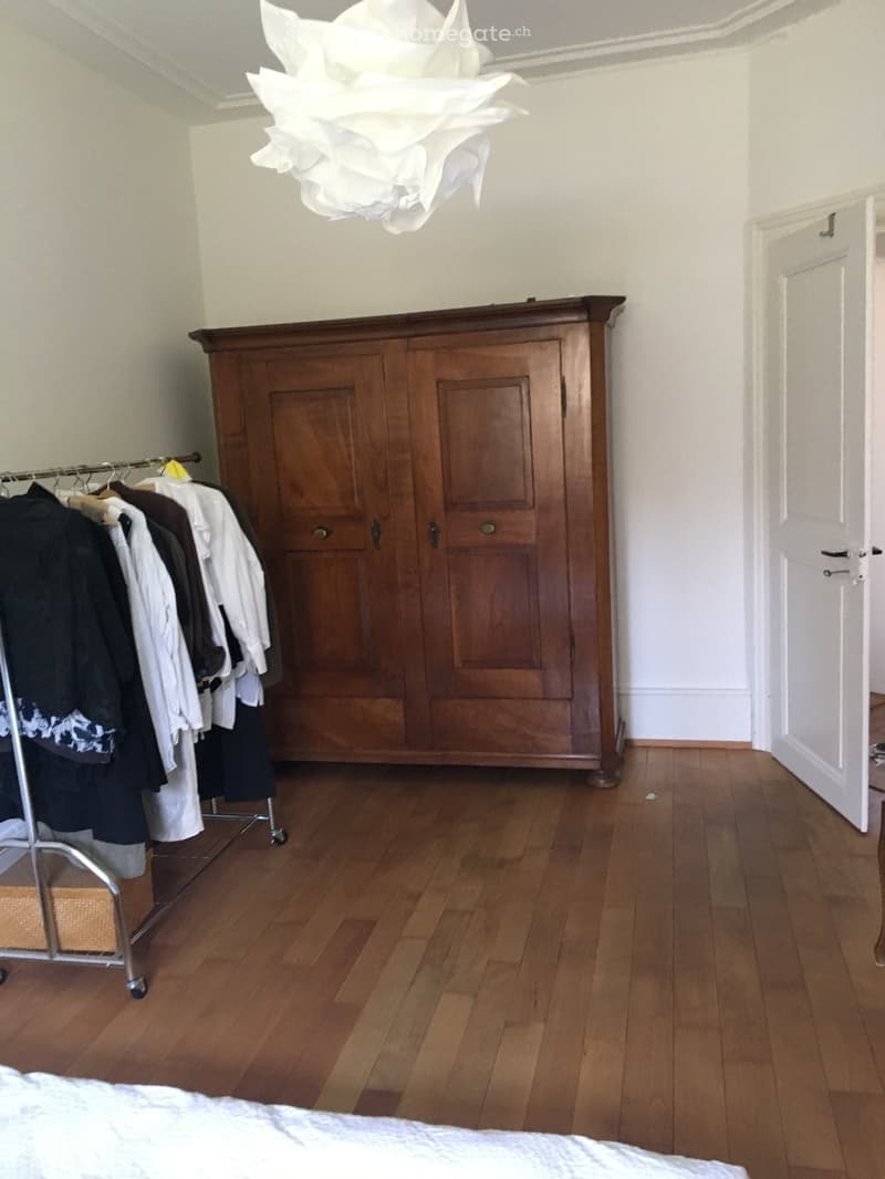 A furnished room with wooden flooring, a large wooden wardrobe, a clothing rack with hanging clothes, and a bed with a patterned bedspread.