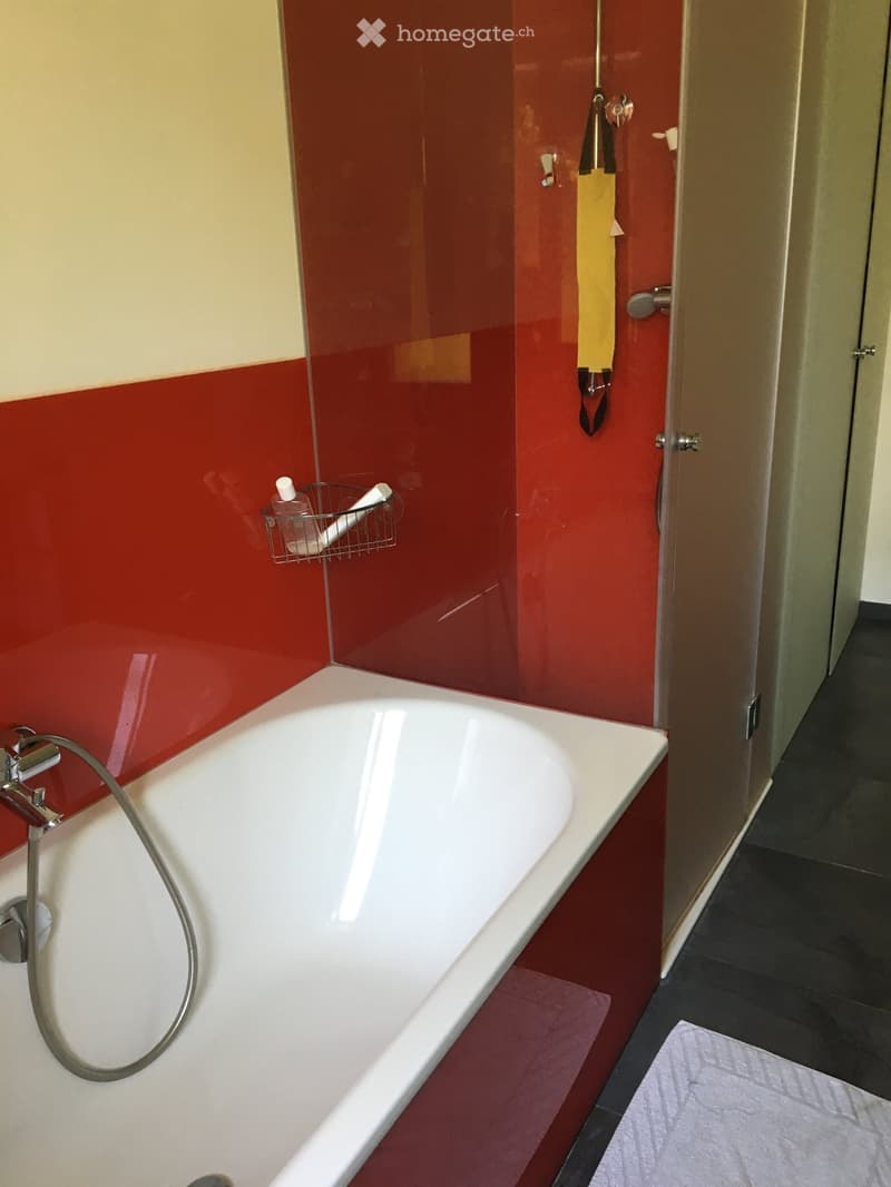 Modern bathroom with a white bathtub, red walls, a shower hose, toothbrush holder, and a partially open bathroom door.