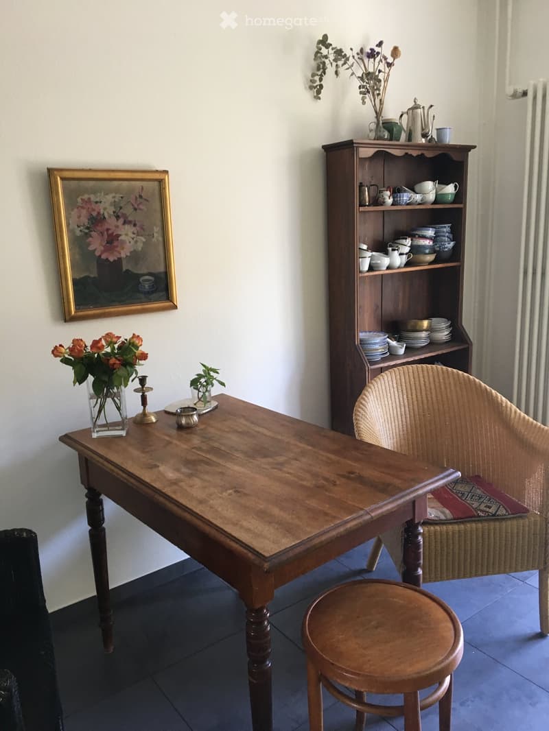 A cozy living room with a wooden table, a wicker chair, a wooden stool, a wooden cupboard filled with dishes and teapots, a vase with flowers, a candle, and a framed painting on the wall.
