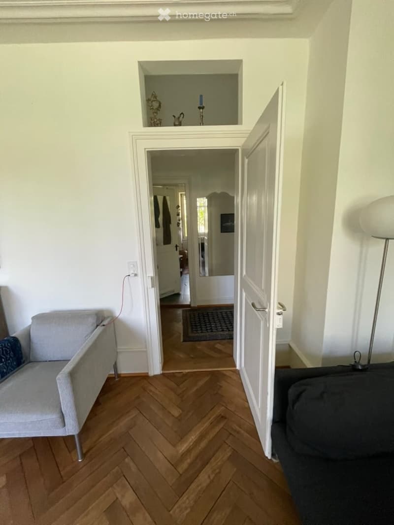 A modern living room with herringbone wooden flooring, white walls, and an open doorway leading to another room. Visible furniture includes a grey sofa, a chair, and a floor lamp.