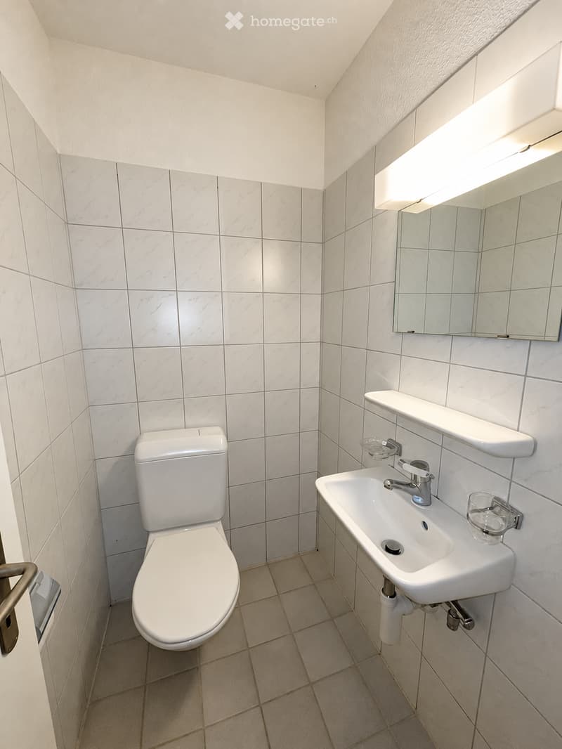 Modern bathroom with white tiled walls and beige tiled floor, featuring a toilet, sink with faucet, and a mirrored shelf above the sink.