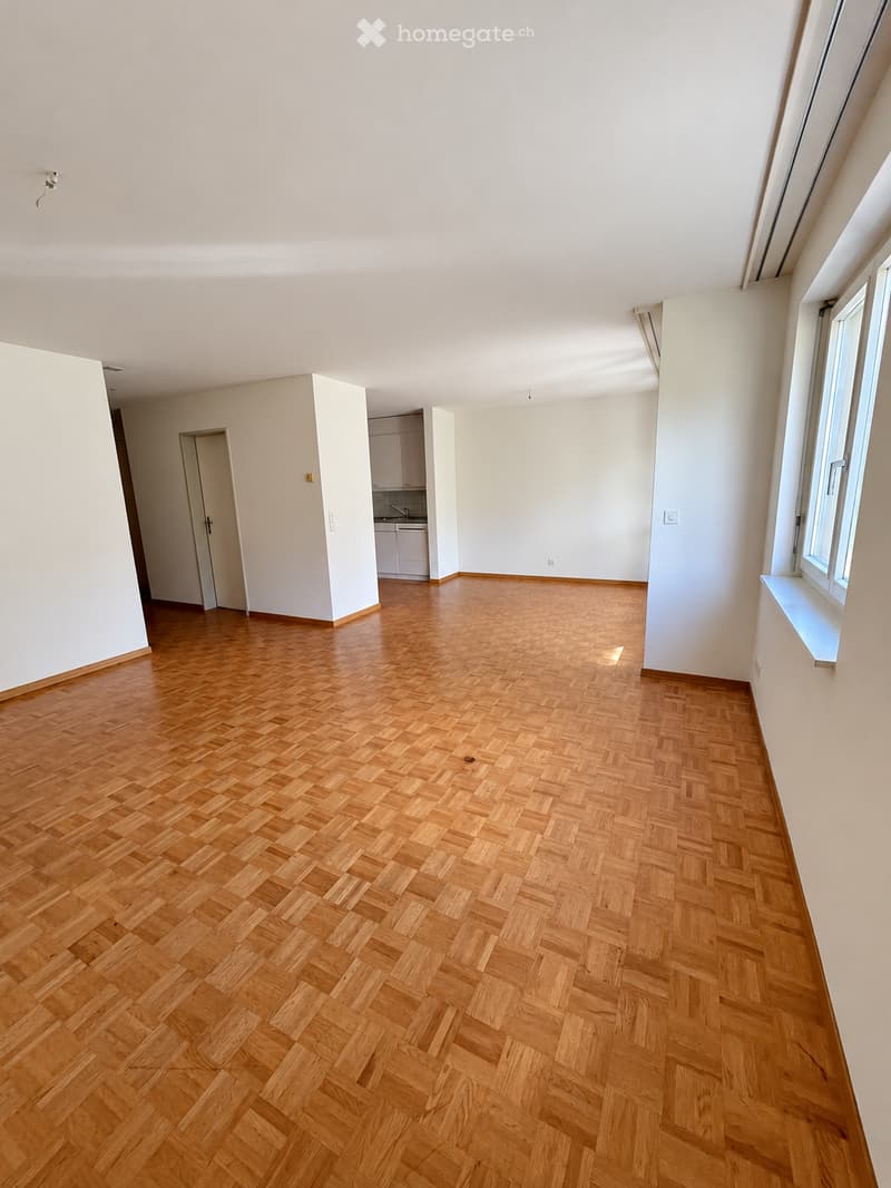 Empty room with wooden herringbone flooring, white walls, and large windows. No furniture or equipment present.