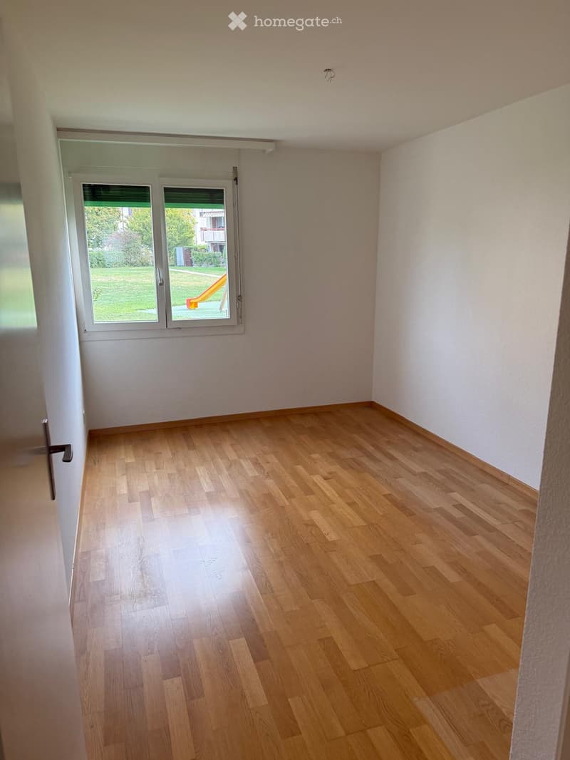 Empty room with light wooden flooring and white walls, featuring a large window with a view of a green outdoor area.
