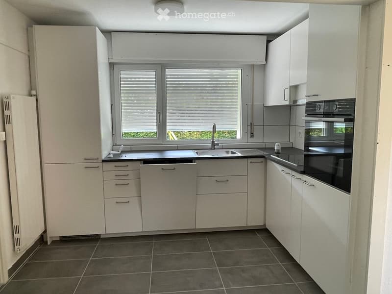 Modern kitchen with white cabinets, dark countertops, and a window with blinds. Includes a sink, faucet, and built-in oven.