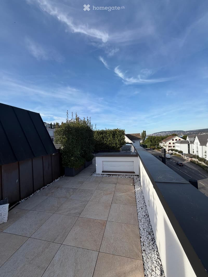 Roof terrace with tiled flooring, low white wall, small white storage unit, and neatly trimmed bushes.