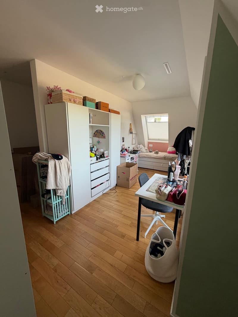 A small, multi-functional room with a bed, desk, and wardrobe. The room features wooden flooring, a slanted ceiling, and a window with blinds.