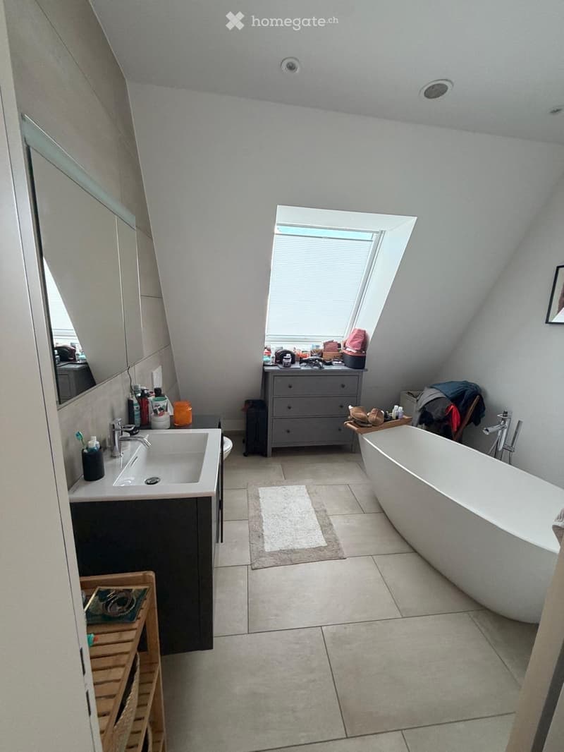 Modern bathroom with a freestanding bathtub, sink, and mirrored cabinet. Features a skylight window, tiled floor, and various toiletries on the counter.