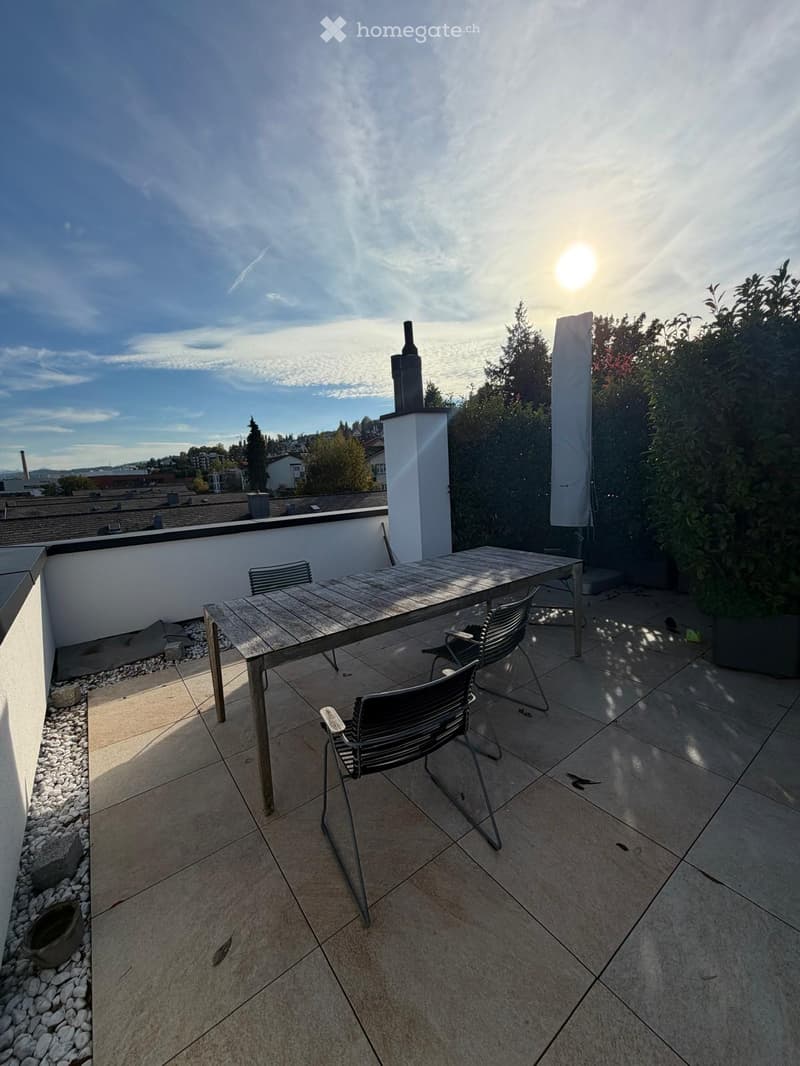 A rooftop terrace with a wooden table and three black chairs, surrounded by a low white wall and greenery.