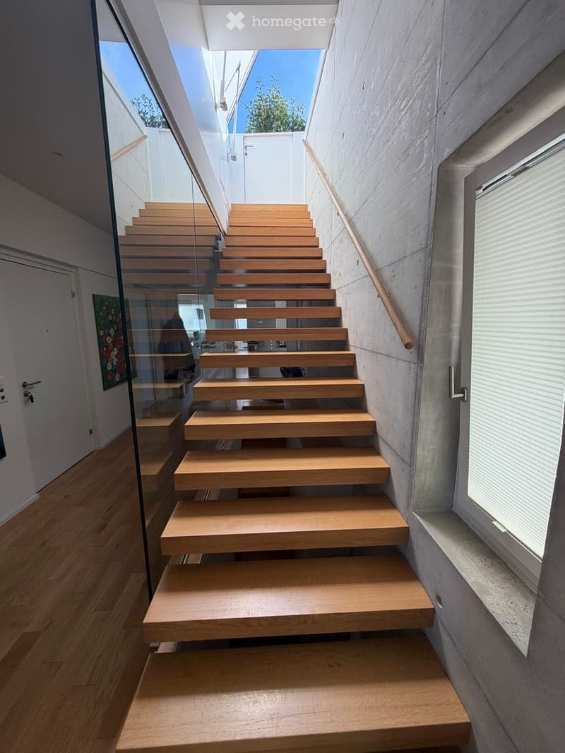 Modern staircase with wooden steps and a glass railing, leading upwards with natural light from the side window.