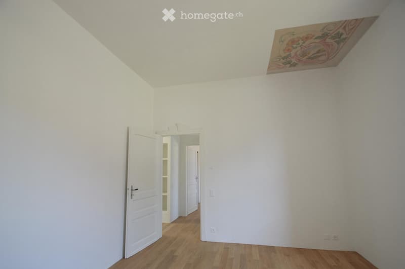 Empty room with white walls, wooden flooring, and a decorative ceiling piece. There is a doorway leading to another room.