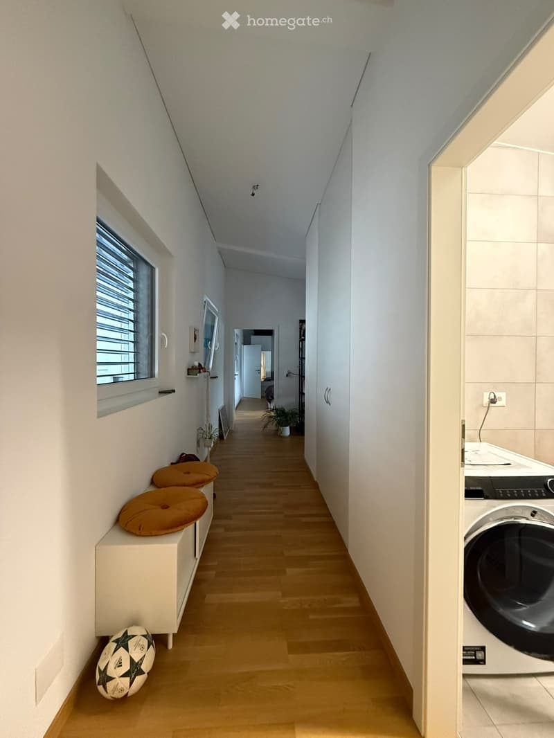 A narrow hallway with light wooden flooring, white walls, and a small window on the left side. There is a bench with two orange cushions and a decorative ball on the floor, and a washing machine is visible at the end of the hallway.