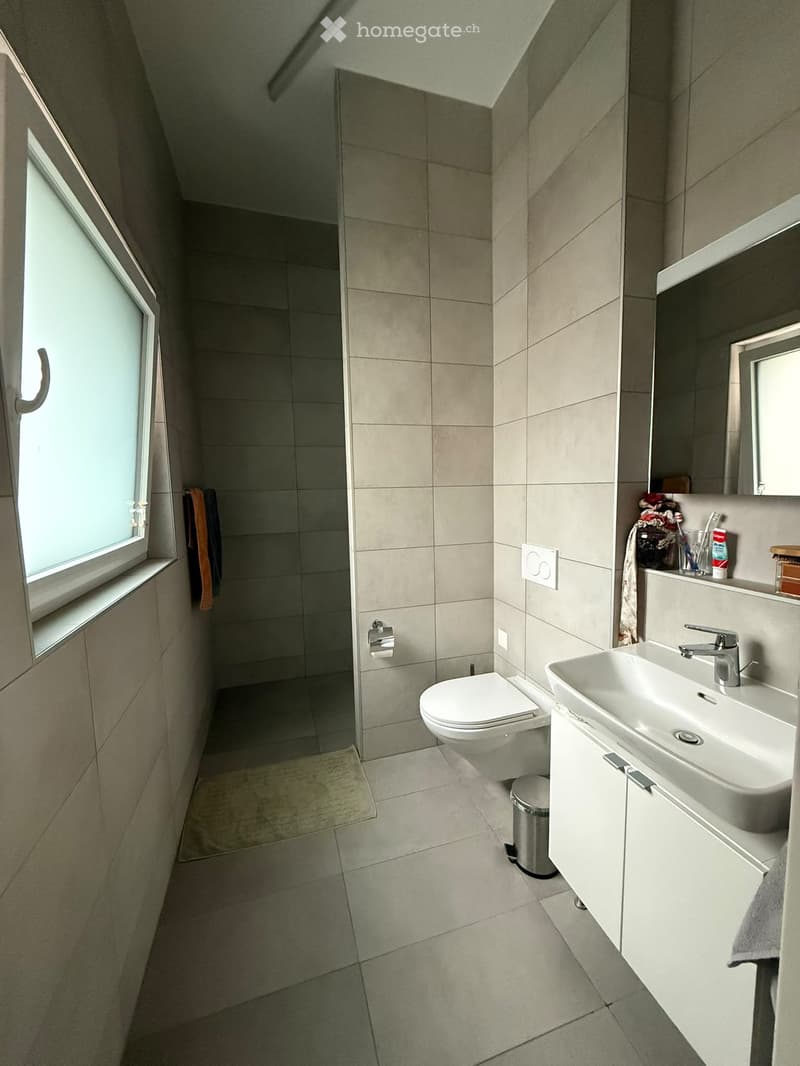 Modern bathroom with beige tiles, a toilet, a sink with a mirror cabinet, and a window with a towel hanging nearby.