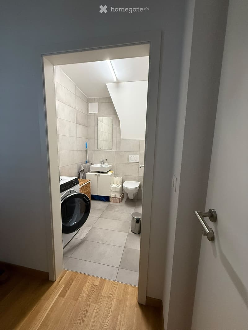 Small bathroom with a toilet, sink, mirror, washing machine, and toilet paper holder. The floor is tiled, and the walls are painted white.