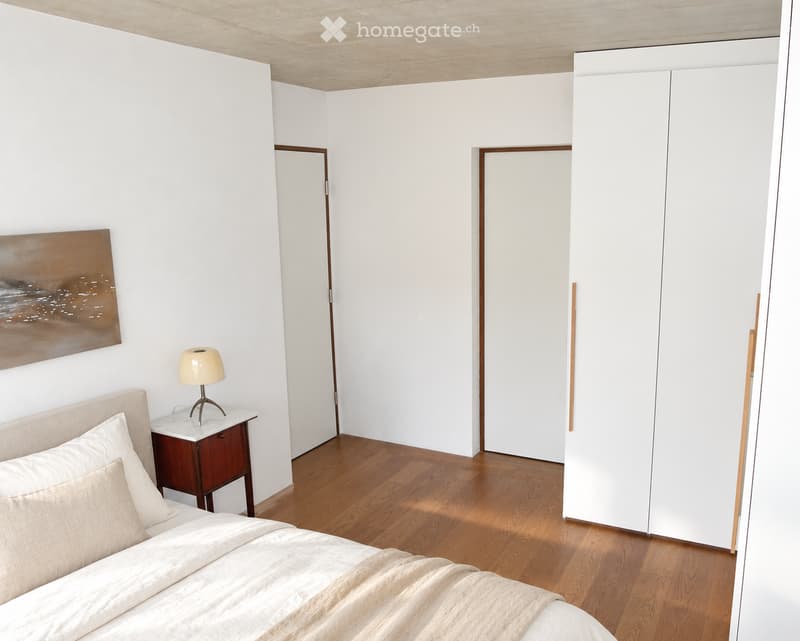 Modern bedroom with a wooden floor, white walls, a large bed with beige bedding, a nightstand with a lamp, and a painting on the wall. There are two doors and a large white wardrobe.