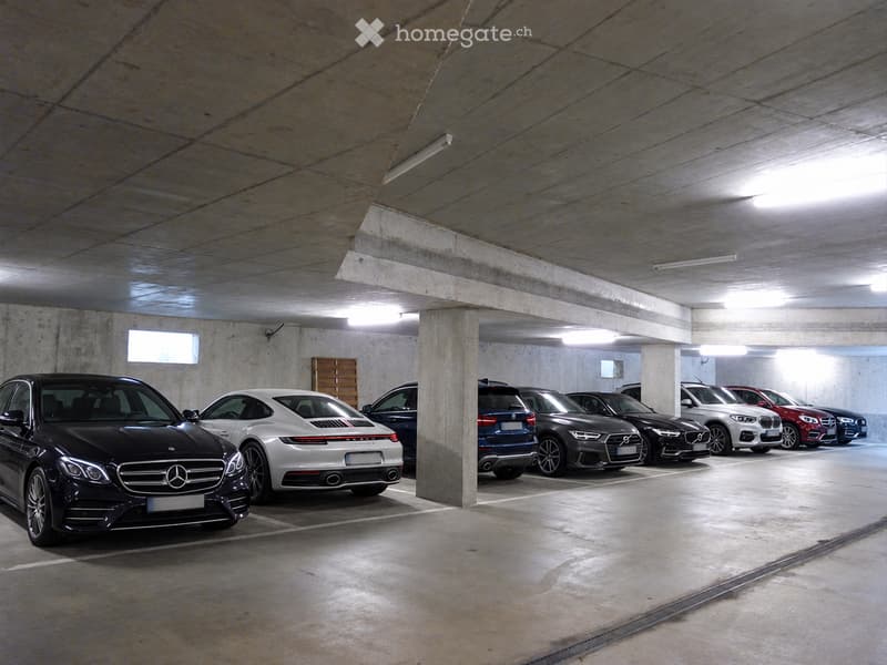 Underground parking area with several parked cars, concrete pillars, and fluorescent lighting.
