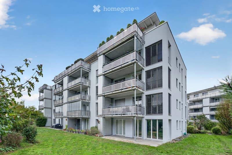 Modern multi-story residential building with white facade, multiple balconies, and surrounding greenery.