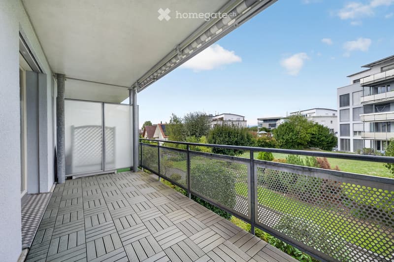 Balcony with wooden decking, metal railing, and a view of a green landscape and modern buildings.