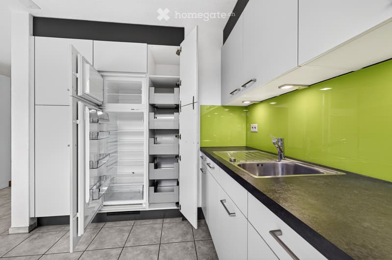 Modern kitchen with white cabinets, stainless steel sink, and open refrigerator. Green accent wall and sleek design.