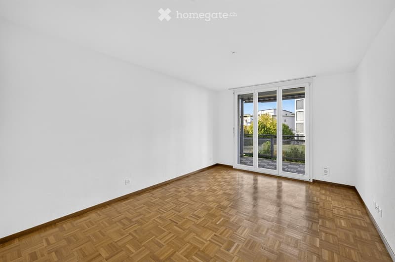 Empty room with white walls and wooden herringbone flooring, featuring large sliding glass doors leading to a balcony.