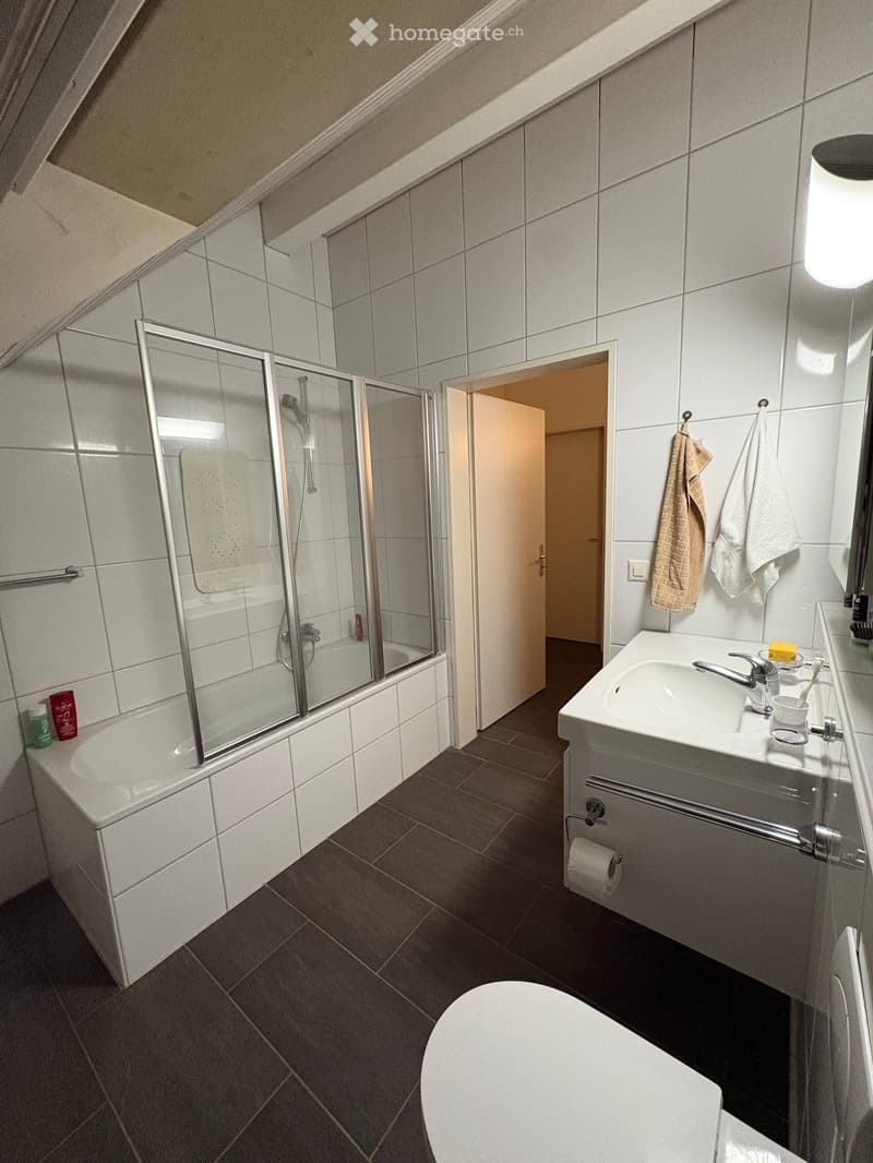 Modern bathroom with white tiles, a glass-enclosed shower, a sink with a faucet, a toilet, and towels hanging on the wall.
