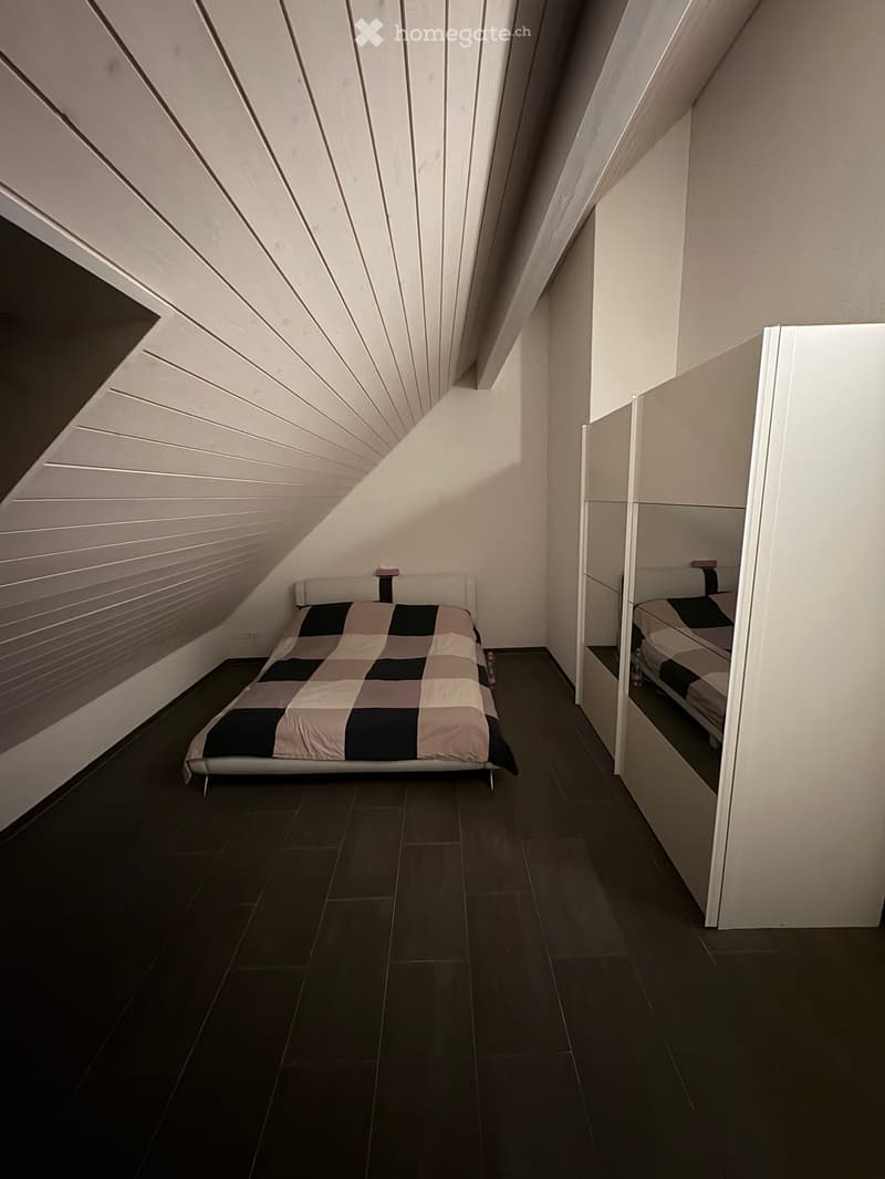 Modern attic bedroom with slanted wooden ceiling, dark tiled floor, and a single bed with a checkered duvet. A mirrored wardrobe is visible on the right side.
