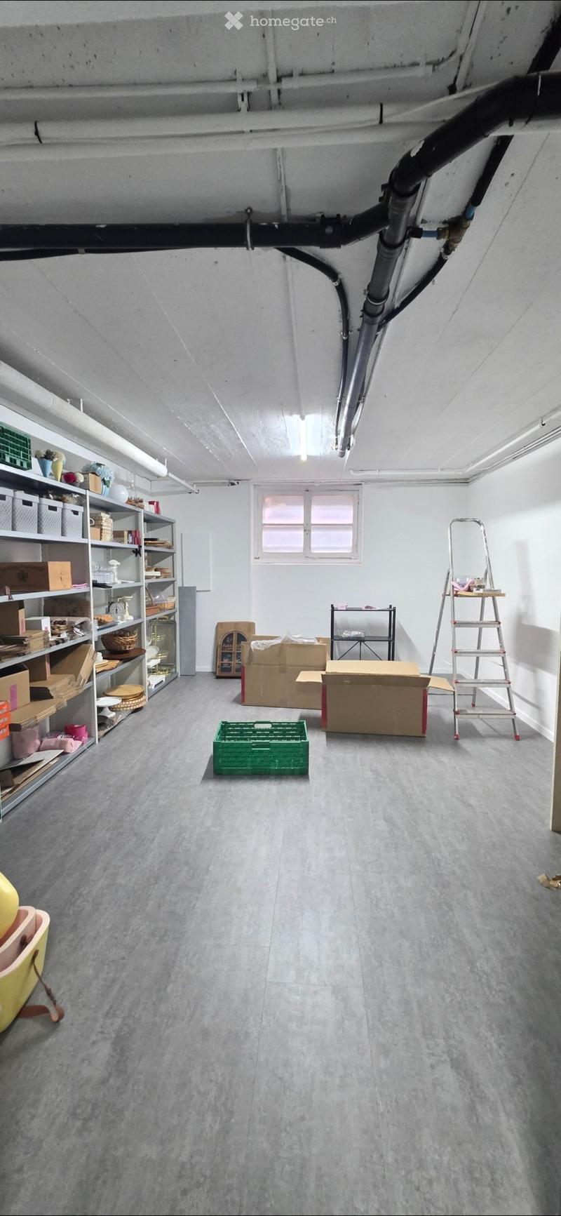 An attic or storage room with exposed pipes and beams, featuring a large window, shelves filled with various items, and several cardboard boxes on the floor.