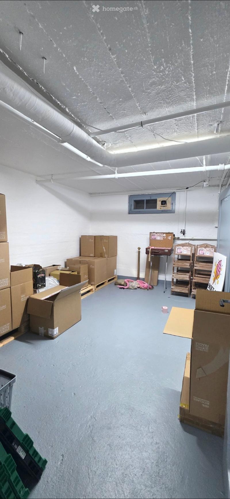 Empty room with bare walls and a concrete floor, featuring stacked cardboard boxes and a small wooden shelf unit.