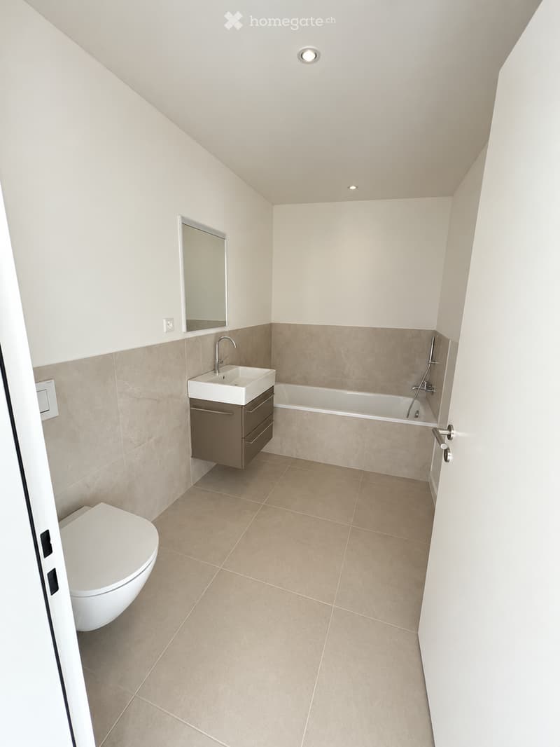 Modern bathroom with beige tiles, a white toilet, a sink with a cabinet, a bathtub, and a mirror on the wall.