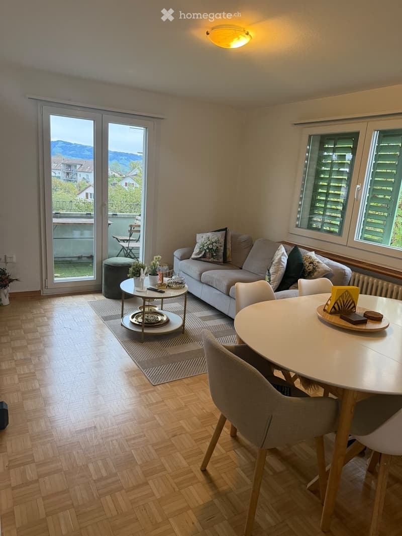 Modern living and dining area with wooden flooring, large windows, and a balcony. Features a grey sofa, coffee table, dining table with chairs, and decorative items.