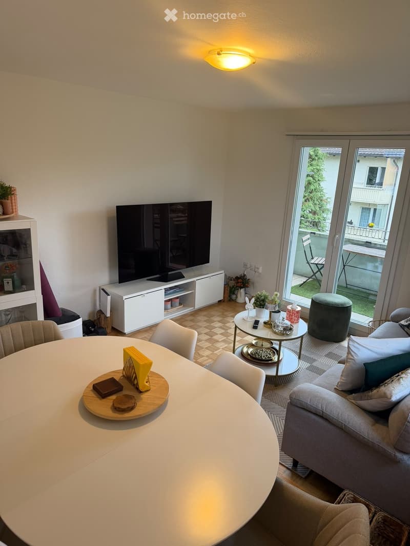 Modern living room with a grey sofa, coffee table, round dining table, chairs, and a television. Features a balcony with outdoor seating visible through large glass doors.