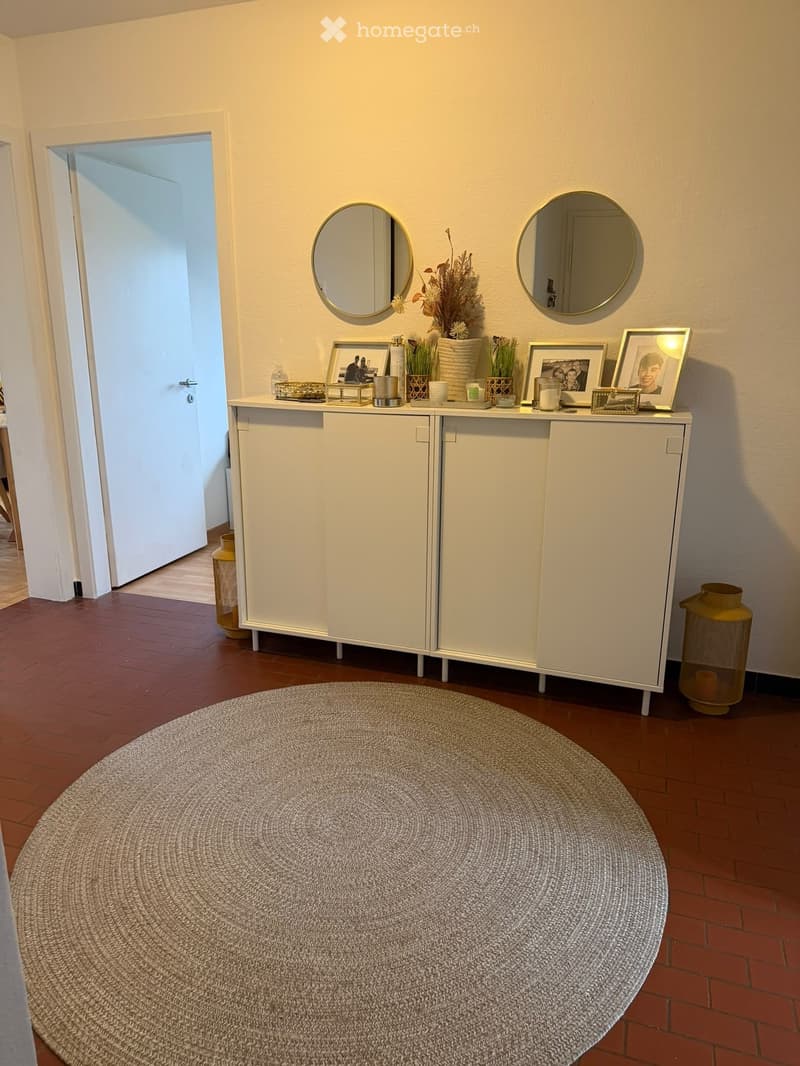 A small, well-lit hallway or entryway with a white cabinet, two round wall mirrors, framed photos, decorative vases, candles, and a round woven rug on a wooden floor.