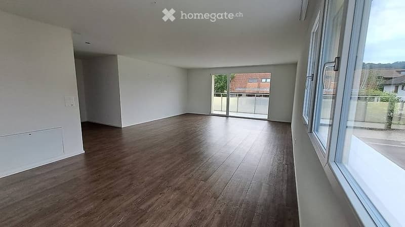 Empty room with wooden flooring, white walls, large windows, and a sliding glass door leading to a balcony.