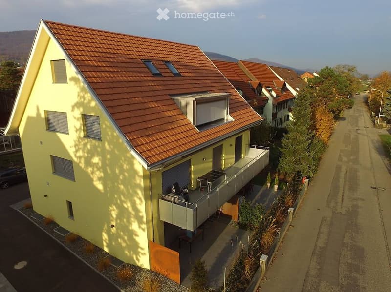 A modern two-story house with a yellow facade and red-tiled roof, featuring a balcony with outdoor furniture and a small garden area.