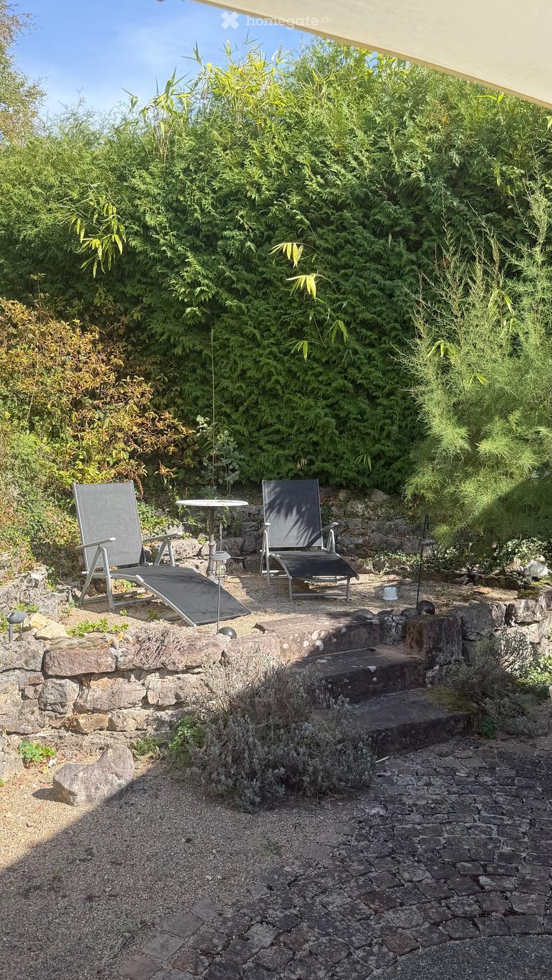 A serene outdoor seating area with two lounge chairs and a small table, surrounded by lush greenery and stone steps leading to a pathway.