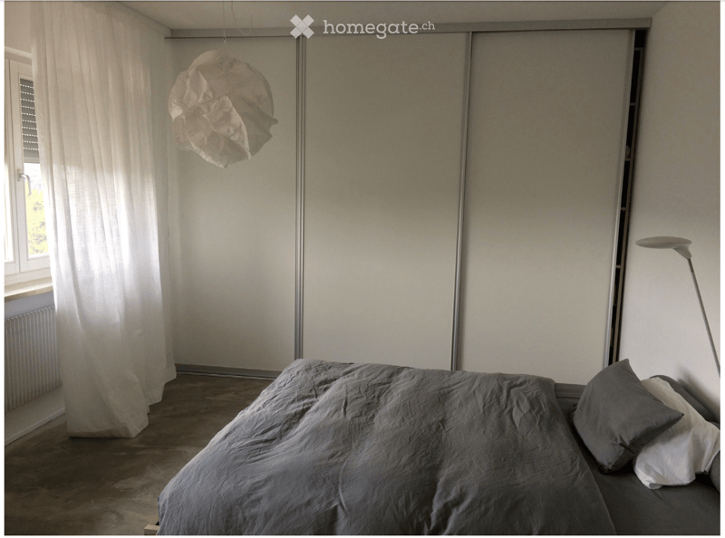 Minimalist bedroom with a grey duvet-covered bed, white curtains, a modern hanging lamp, and a sleek floor lamp near the window.