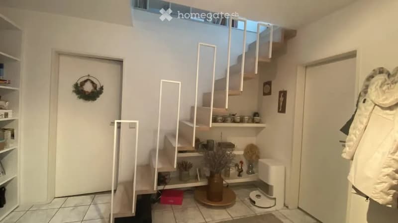 Modern hallway with white walls, tiled floor, staircase leading to an upper level, decorative items including a wreath on the door, small shelves with various items, and a small round table with a vase of dried flowers.