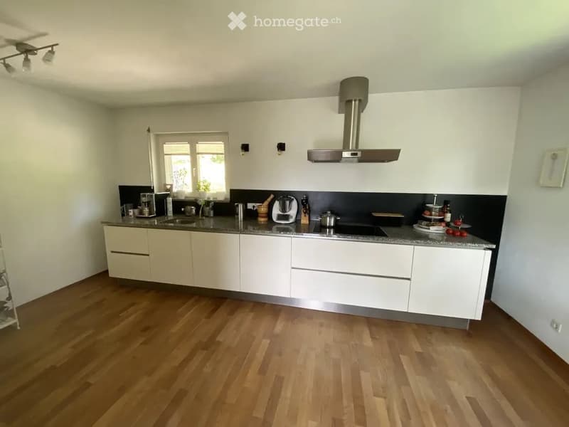 Modern kitchen with white cabinets, wooden flooring, and stainless steel appliances including a stove, oven, and coffee maker. The countertop features various kitchen items such as a knife block, dish rack, and small appliances.