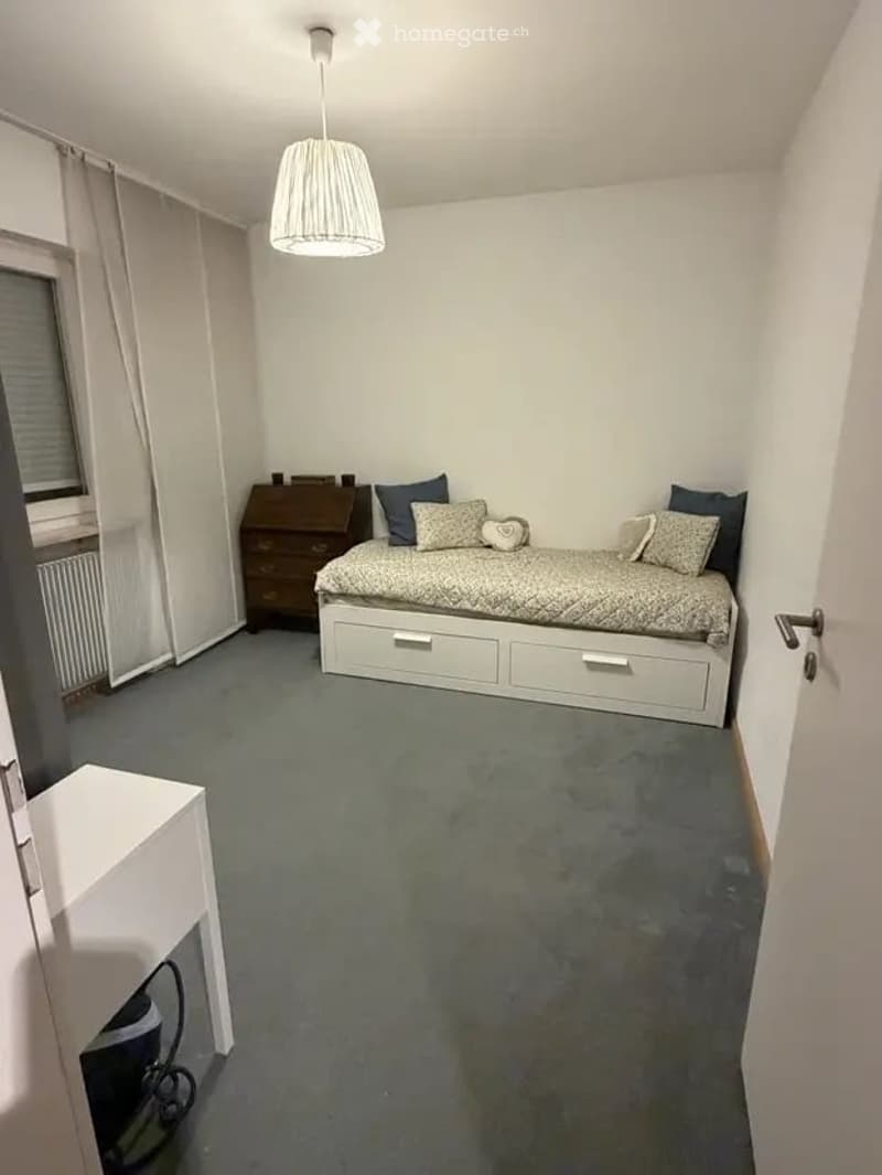 Cozy bedroom with a single bed, nightstand, pillows, and a drawer unit. The room features a grey carpet, white walls, and a hanging lamp.