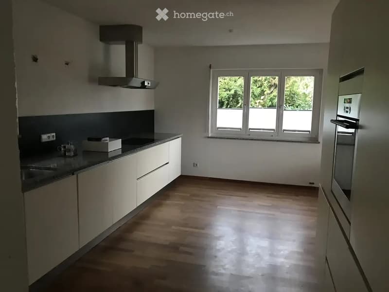 Modern kitchen with light-colored cabinets, a stainless steel oven, a range hood, and a dark countertop. The floor is wooden, and there is a large window with a view of greenery.
