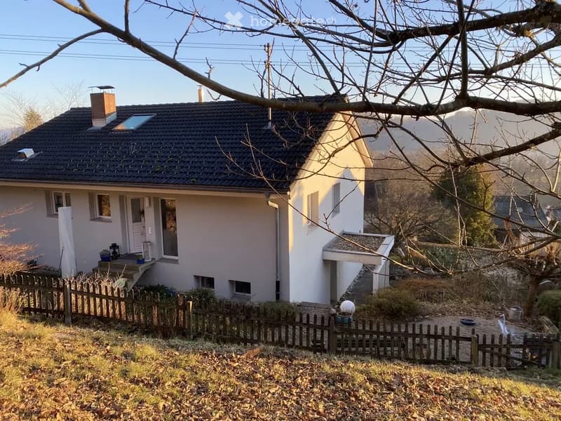 Single-story house with a dark roof, white walls, and a small porch. The house has a wooden fence surrounding the front yard, with some plants and a small garden area visible.