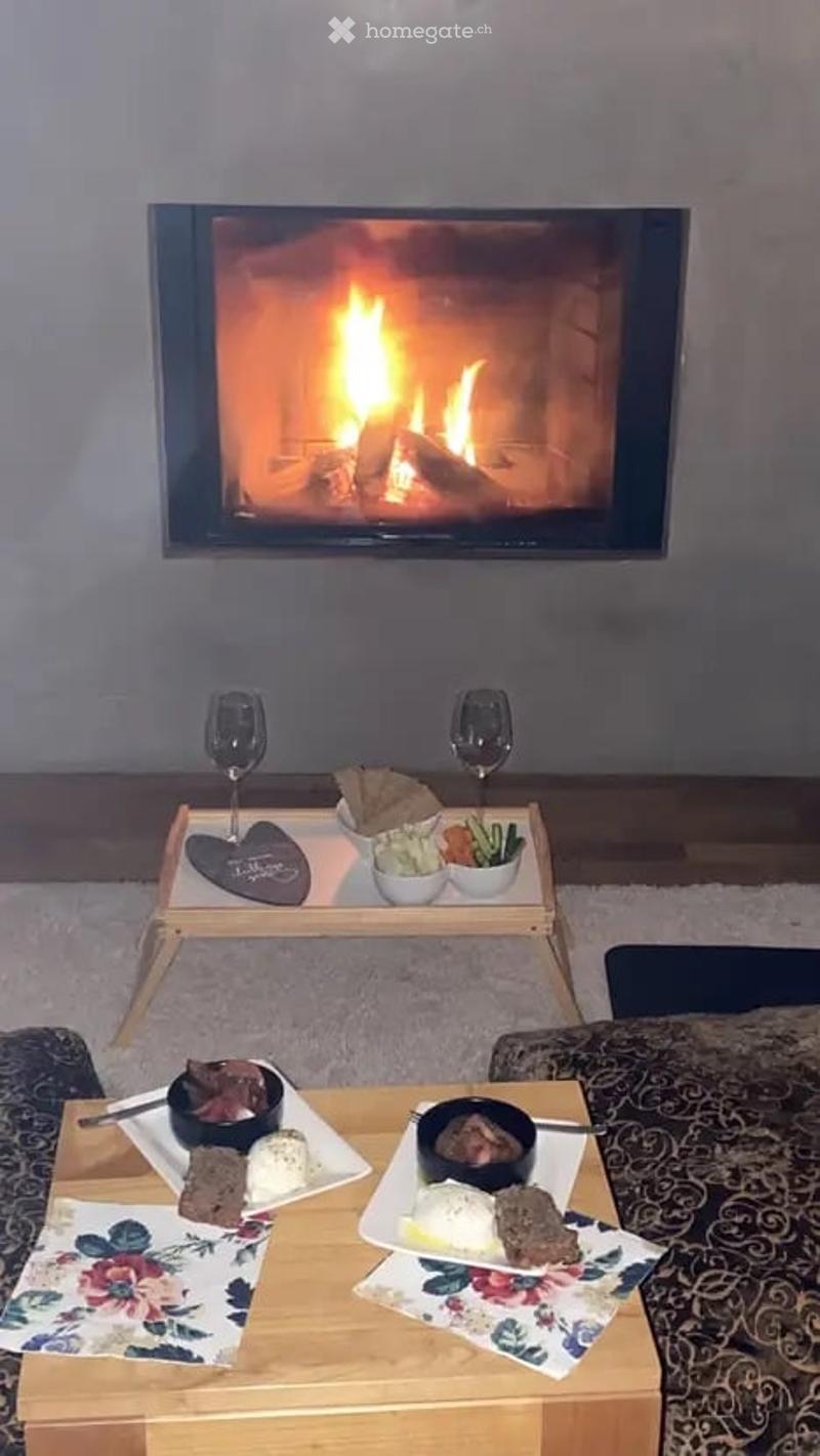 A cozy living room with a modern fireplace, a coffee table with wine glasses and snacks, and a dining table set with plates of food and drinks.