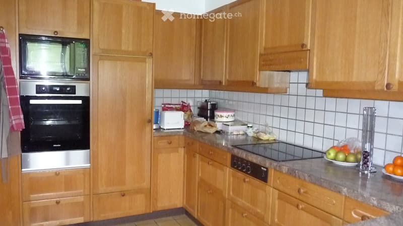 The kitchen features wooden cabinets, a stainless steel oven, microwave, toaster, and induction stove. The countertop has a fruit bowl with oranges, apples, and other items. The walls are tiled, and there is a dish rack visible.